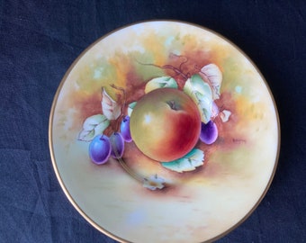 May include: A decorative plate featuring a hand-painted still life of a ripe apple and several purple plums. The fruit is surrounded by green leaves and brown vines, set against a cream-colored background with a gold rim.