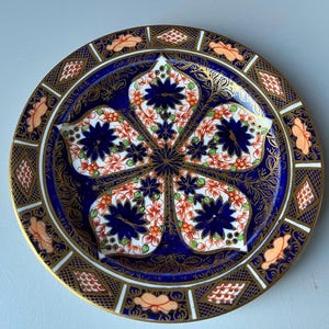 May include: A decorative plate with a blue and gold floral design. The plate features a central floral pattern with orange and white accents, surrounded by a gold border with geometric and floral details. The plate is likely made of porcelain.