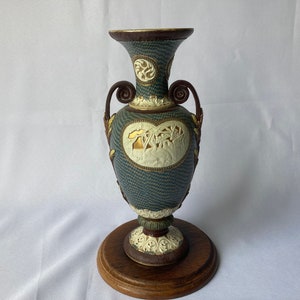 May include: An ornate, antique vase with a dark teal textured body, brown handles, and a decorative base. The vase features cream-colored relief carvings and gold accents. It sits on a round, wooden base, showcasing a classic, vintage design.