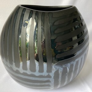 Può includere: Un vaso in ceramica grigio scuro con finitura lucida. Il vaso ha una forma arrotondata con un'ampia apertura ed è decorato con strisce verticali e orizzontali in varie tonalità di grigio e bianco. Il vaso è alto circa 25 cm.