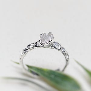 May include: A silver ring with a rough, clear diamond set in a textured band.