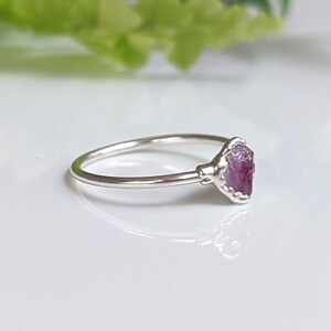 May include: A silver ring with a raw, pink gemstone. The gemstone is set in a decorative bezel, and the ring band is thin and polished. The ring is shown on a white surface, with a blurred green background. The ring is likely made of sterling silver.