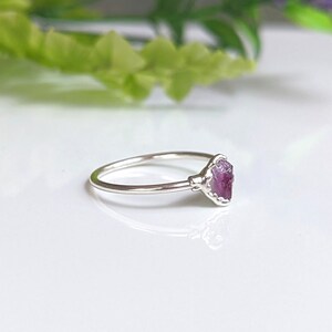 May include: A silver ring with a raw, pink gemstone. The gemstone is set in a detailed bezel setting, and the ring band is thin and polished. The ring is displayed on a white surface with soft, blurred greenery in the background.