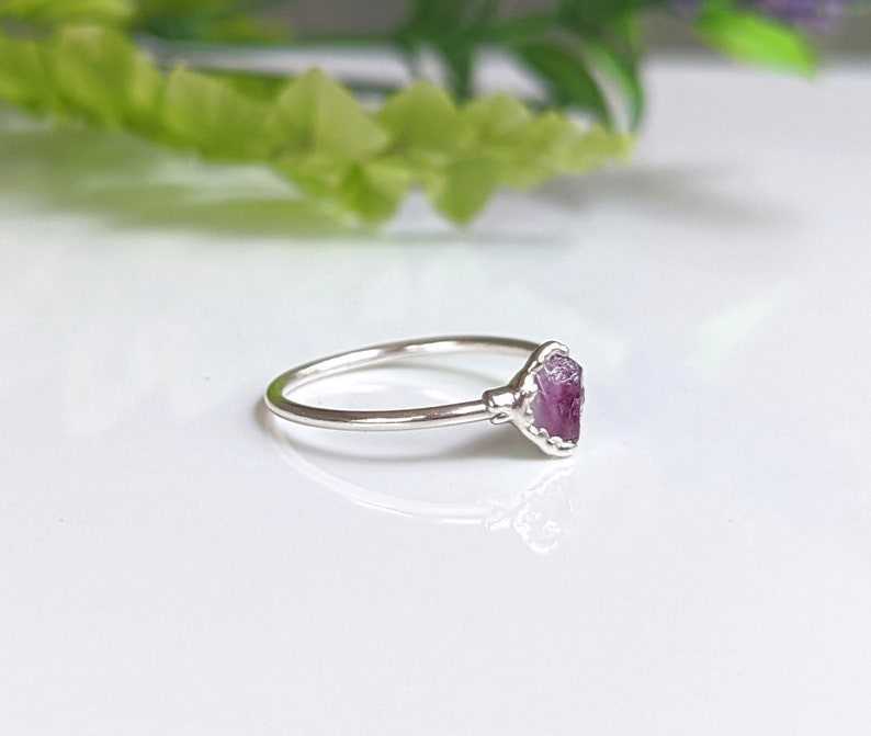 May include: A silver ring with a raw, pink gemstone. The gemstone is set in a detailed bezel setting, and the ring band is thin and polished. The ring is displayed on a white surface with soft, blurred greenery in the background.