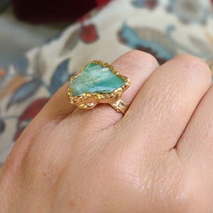 A hand wearing a Solid 14k Gold ring featuring a large raw and uncut Aqua blue Peruvian Opal