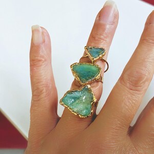 A hand wearing 3 different sizes of Solid 14k Gold ring featuring a large raw and uncut Aqua blue Peruvian Opal
