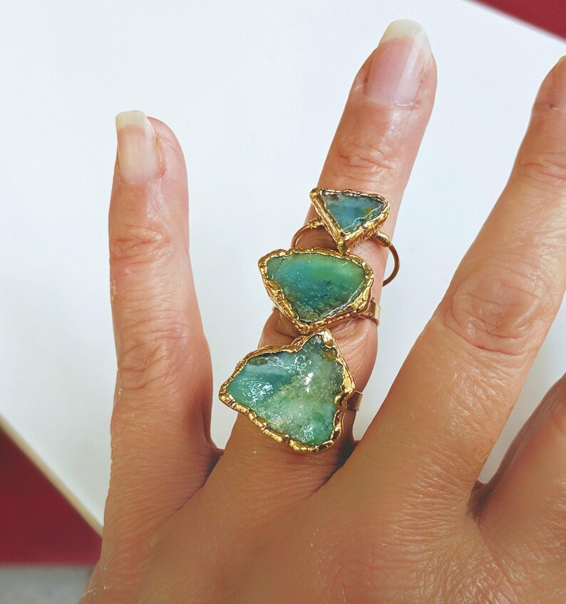 A hand wearing 3 different sizes of Solid 14k Gold ring featuring a large raw and uncut Aqua blue Peruvian Opal