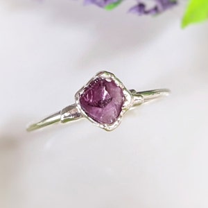 May include: A delicate silver ring featuring a raw, square-cut, deep purple gemstone. The gemstone is set in a textured bezel, and the band is thin and polished. The ring is a unique piece of jewellery.