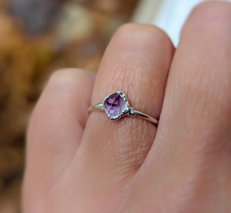 May include: A silver ring with a raw, faceted amethyst gemstone. The purple gemstone is set in a delicate, handcrafted bezel. The ring's band is thin, emphasising the unique shape and colour of the amethyst. A piece of jewellery.