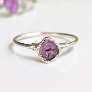 May include: A silver ring with a raw amethyst gemstone. The purple gemstone is set in a textured bezel, with a delicate band. The ring is a unique piece of jewellery.