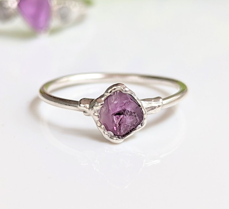 May include: A silver ring with a raw amethyst gemstone. The purple gemstone is set in a textured bezel, with a delicate band. The ring is a unique piece of jewellery.