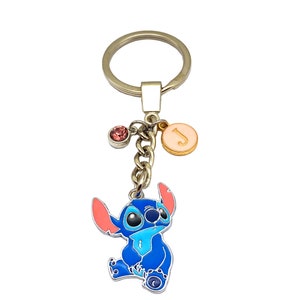May include: Blue Stitch character keychain with a pink and gold charm with the letter J and a pink crystal charm. The keychain is attached to a silver key ring.
