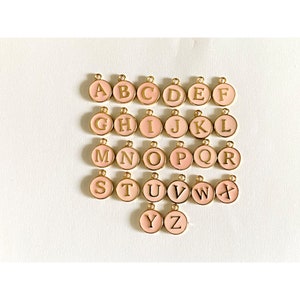May include: A set of 26 gold-colored alphabet charms with pink enamel. Each charm features a letter of the alphabet in a gold-colored font.