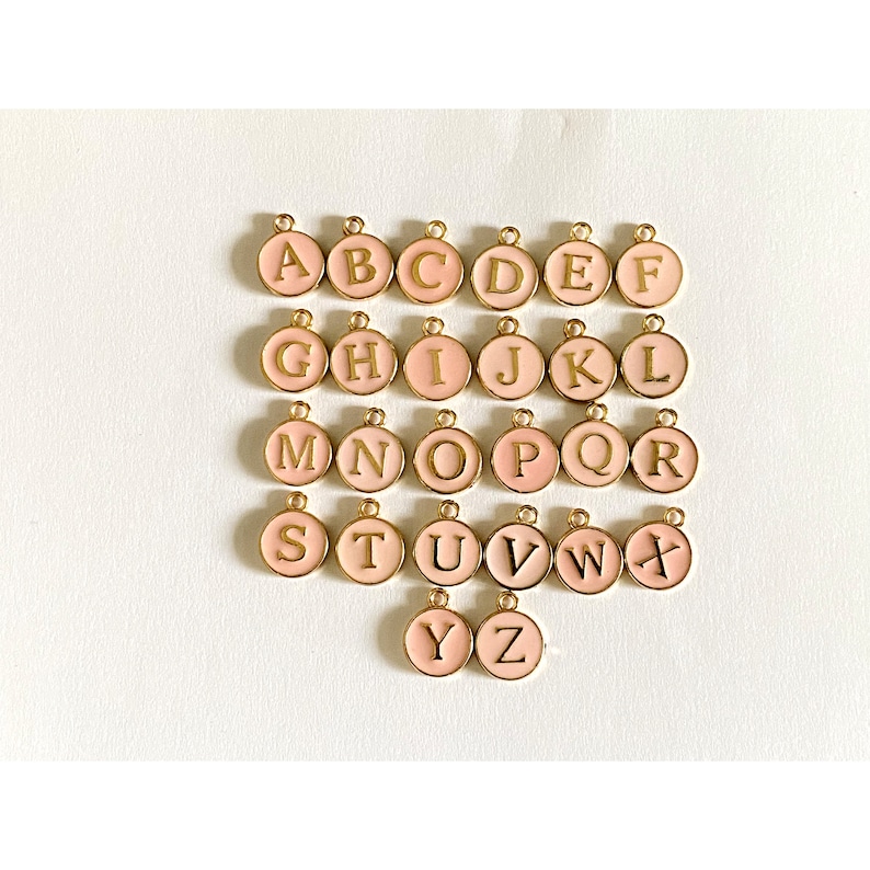 May include: A set of 26 gold-colored alphabet charms with pink enamel. Each charm features a letter of the alphabet in a gold-colored font.