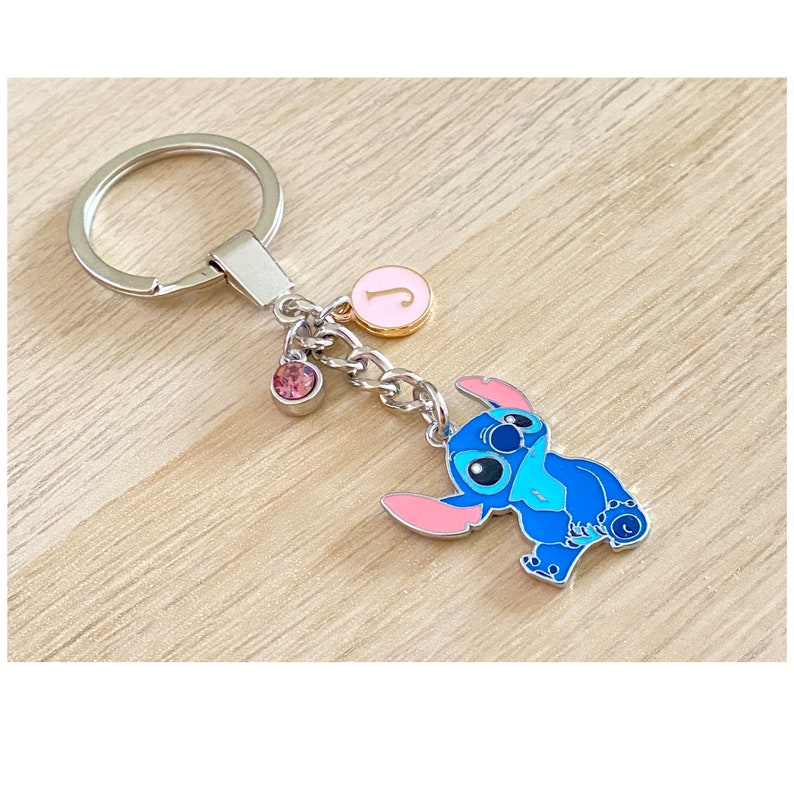 May include: A blue Stitch character keychain with pink ears and a pink and gold charm with the letter J. The keychain is attached to a silver key ring.