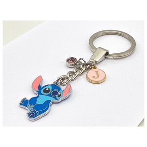 May include: Blue Stitch character keychain with a pink and gold charm with the letter J. The keychain also has a pink rhinestone charm.