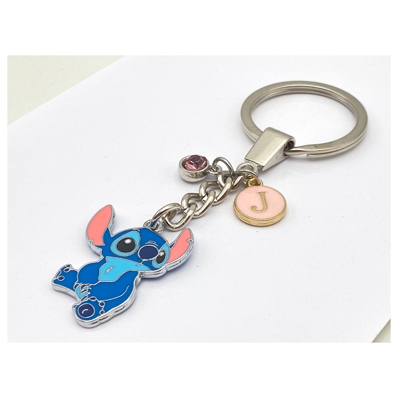 May include: Blue Stitch character keychain with a pink and gold charm with the letter J. The keychain also has a pink rhinestone charm.