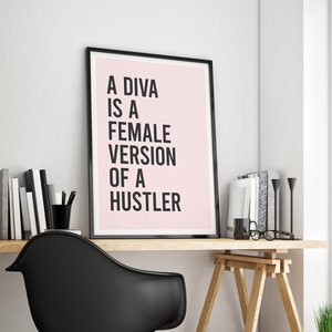 May include: A black framed poster with a pink background and white text that reads "A DIVA IS A FEMALE VERSION OF A HUSTLER". The poster is hanging on a white wall above a wooden desk with a black chair and a wooden easel.