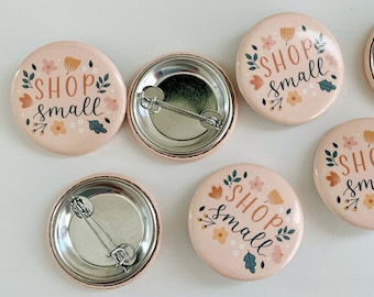 Könnte beinhalten: Sechs rosa Anstecknadeln mit dem Text "Shop Small" in schwarzer Schrift, umgeben von floralen Designs. Die Knöpfe haben eine silberne Rückseite mit einer Sicherheitsnadel.