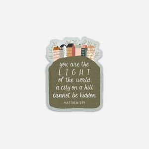 Op de afbeelding: Een sticker met een groene achtergrond en een citaat uit de Bijbel, Mattheüs 5:14, "Gij zijt het licht der wereld. Een stad op een heuvel kan niet verborgen worden." De sticker toont een fantasievolle illustratie van een stad met kleurrijke huizen.