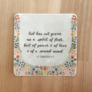 Op de afbeelding: Een witte onderzetter met een bloemige rand en een citaat uit 2 Timoteüs 1:7: "Want God heeft ons niet de geest van angst gegeven, maar de geest van kracht, liefde en bezonnenheid."