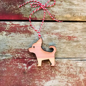 Puede incluir: Adorno de chihuahua de madera con acabado de madera natural. El adorno con forma de perro está suspendido por una cuerda de hilo rojo y blanco atada en un lazo. El adorno está sobre un fondo de madera rústica y desgastada.