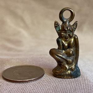 May include: A small, bronze-colored figurine of a seated mythical creature with pointed ears and a loop at the top. The figurine is next to a quarter coin for size comparison. The creature appears to be holding a small object.