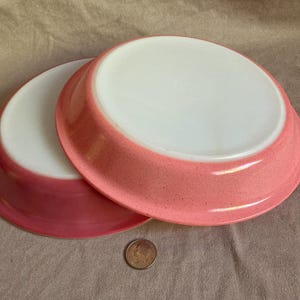 May include: Two round pie dishes with white interiors and speckled pink exteriors. The dishes are stacked slightly offset, revealing the smooth white base and the textured pink rim. A small circular metal coin is placed in front for scale.