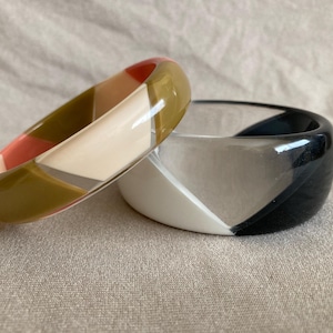 May include: Two colorful bangles. One is a mix of olive green, peach, and cream geometric shapes. The other is black, white, and gray. Both are made of a glossy material and rest on a neutral fabric surface.