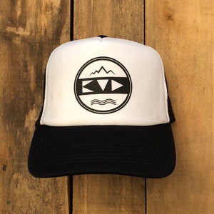 Puede incluir: Una gorra de camionero blanca y negra con un logotipo circular negro que presenta una cordillera, un triángulo y las letras "KVD" con líneas onduladas debajo.