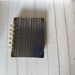 May include: A small spiral-bound notebook featuring a dark blue cover with a repeating gold-colored herringbone pattern. The notebook has gold-toned metal wire binding along the left side and gold-colored metal corner protectors on the cover.