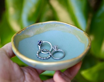 May include: A small, light blue ceramic ring dish with a gold rim. The dish is holding three silver rings, one with a blue stone.