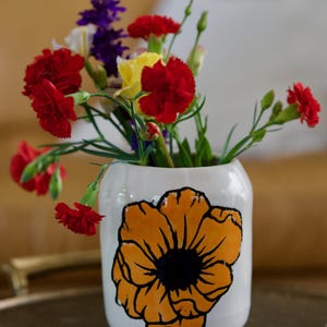 May include: A white ceramic vase with a hand-painted orange flower design, filled with a vibrant arrangement of red carnations, yellow flowers, and purple wildflowers. The vase sits on a brown tray.