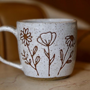 May include: A white ceramic mug with a speckled finish and a brown handle. The exterior features a hand-drawn brown line art design of three distinct wildflowers with stems and leaves. The base of the mug is a solid brown, unglazed ceramic material.