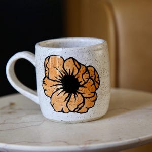 May include: A ceramic mug with a speckled white finish and a single handle. The front of the mug features a hand-painted illustration of a large, orange flower with black outlines and a solid black center, creating a bold contrast against the light base.