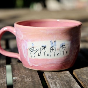 May include: A pink ceramic mug with a handle. The mug features the word "LOVE" spelled out in American Sign Language on a white rectangular patch. The mug is sitting on a wooden surface.