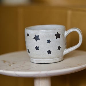 May include: A ceramic mug with a white speckled finish and black star designs. The mug has a curved handle and sits on a small, round, marble-like table. The mug is approximately 4 inches tall.