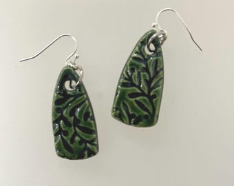 May include: A pair of green ceramic earrings with a leaf design. The earrings are teardrop-shaped with a dark green leaf pattern and silver-colored ear wires. The earrings are hanging against a white background.