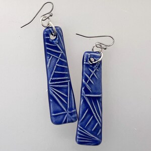 May include: A pair of cobalt blue ceramic earrings with a geometric, etched design. The earrings are rectangular with rounded edges and silver-colored ear wires. The etched pattern is white and creates a contrasting texture.