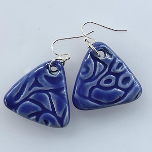 May include: A pair of blue ceramic earrings with a triangular shape. The earrings have a glossy finish and a white, swirling pattern. The ear wires are silver-colored.