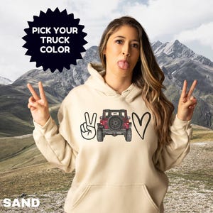May include: A sand-colored hooded sweatshirt featuring a graphic print on the front. The design includes a hand making a peace sign, a red off-road truck viewed from the rear, and a heart shape. The text PICK YOUR TRUCK COLOR is visible.