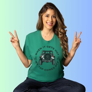 Peut inclure: Un t-shirt vert &agrave; manches courtes avec un motif repr&eacute;sentant l'arri&egrave;re d'un v&eacute;hicule tout-terrain blanc avec une roue de secours. Le texte indique "WHEN IT GETS HOT MY TOP COMES OFF" avec deux ic&ocirc;nes de signe de paix sur les c&ocirc;t&eacute;s.