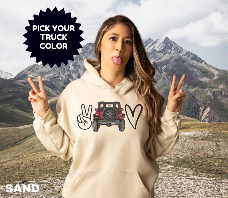 May include: A sand-colored hooded sweatshirt featuring a graphic print on the front. The design includes a hand making a peace sign, a red off-road truck viewed from the rear, and a heart shape. The text PICK YOUR TRUCK COLOR is visible.