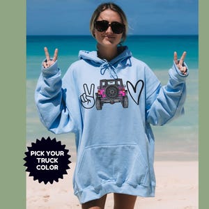 May include: A light blue hooded sweatshirt featuring a graphic print of a pink off-road vehicle positioned between a peace sign and a heart symbol. The text "PICK YOUR TRUCK COLOR" is displayed in a black starburst shape on the lower left side.