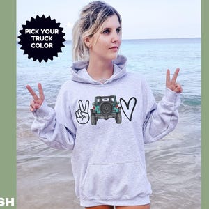 May include: A light gray hooded sweatshirt featuring a graphic print on the chest. The design includes a peace sign, a teal-colored off-road truck viewed from the rear, and a heart shape. The text PICK YOUR TRUCK COLOR is printed above.