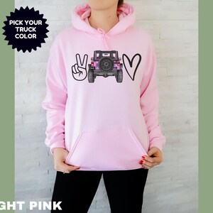 May include: A light pink hooded sweatshirt featuring a graphic print on the chest. The design includes a black peace sign, a black outline of a heart, and a central illustration of a pink off-road vehicle. The text PICK YOUR TRUCK COLOR is visible.