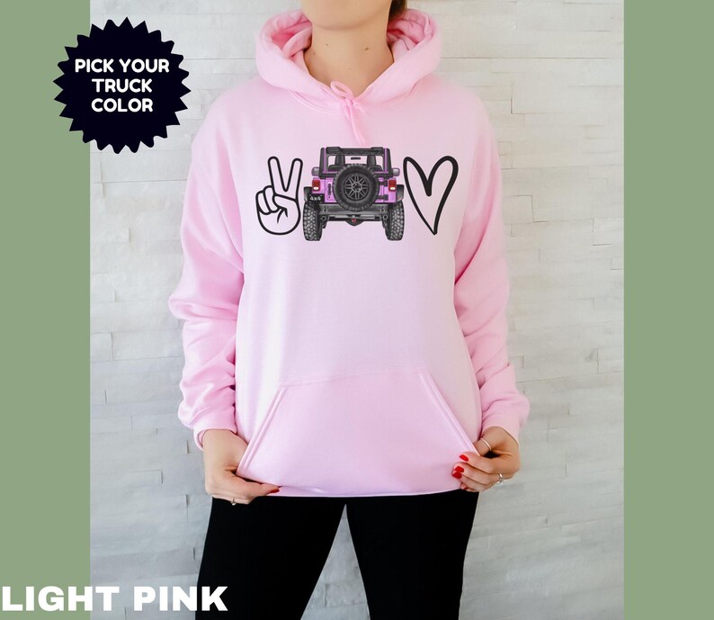 May include: A light pink hooded sweatshirt featuring a graphic print on the chest. The design includes a black peace sign, a black outline of a heart, and a central illustration of a pink off-road vehicle. The text PICK YOUR TRUCK COLOR is visible.