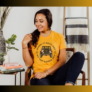 Peut inclure: Un t-shirt jaune moutarde &agrave; manches courtes avec un motif repr&eacute;sentant l'arri&egrave;re d'un v&eacute;hicule tout-terrain noir. Le texte "WHEN IT GETS HOT MY TOP COMES OFF" est imprim&eacute; en arc de cercle autour du v&eacute;hicule, avec deux ic&ocirc;nes de signe de paix.