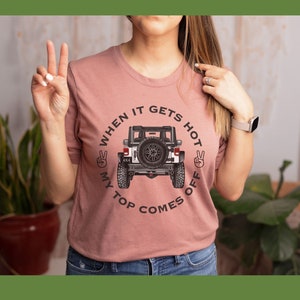 Peut inclure: Un t-shirt &agrave; manches courtes rose poudr&eacute; avec un motif repr&eacute;sentant un v&eacute;hicule tout-terrain blanc vu de l'arri&egrave;re. Le texte "WHEN IT GETS HOT MY TOP COMES OFF" est imprim&eacute; en noir autour du v&eacute;hicule, avec deux ic&ocirc;nes de signe de paix.