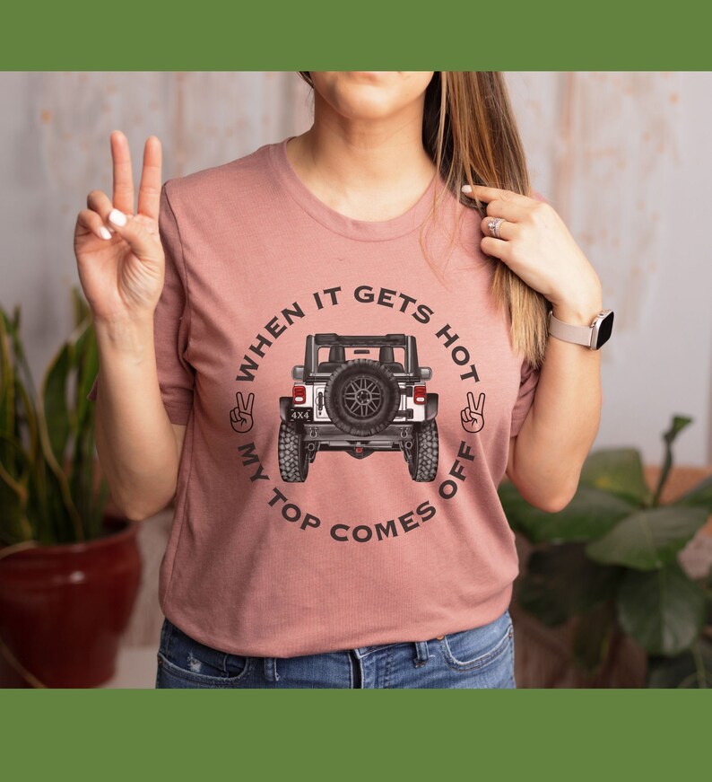 Peut inclure: Un t-shirt &agrave; manches courtes rose poudr&eacute; avec un motif repr&eacute;sentant un v&eacute;hicule tout-terrain blanc vu de l'arri&egrave;re. Le texte "WHEN IT GETS HOT MY TOP COMES OFF" est imprim&eacute; en noir autour du v&eacute;hicule, avec deux ic&ocirc;nes de signe de paix.